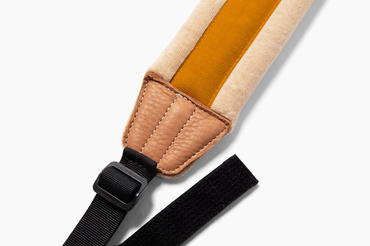 Close-up of a shoulder strap with a beige fabric body, a mustard yellow center panel, tan leather padding, and a black adjustable strap with a plastic buckle on a white background.