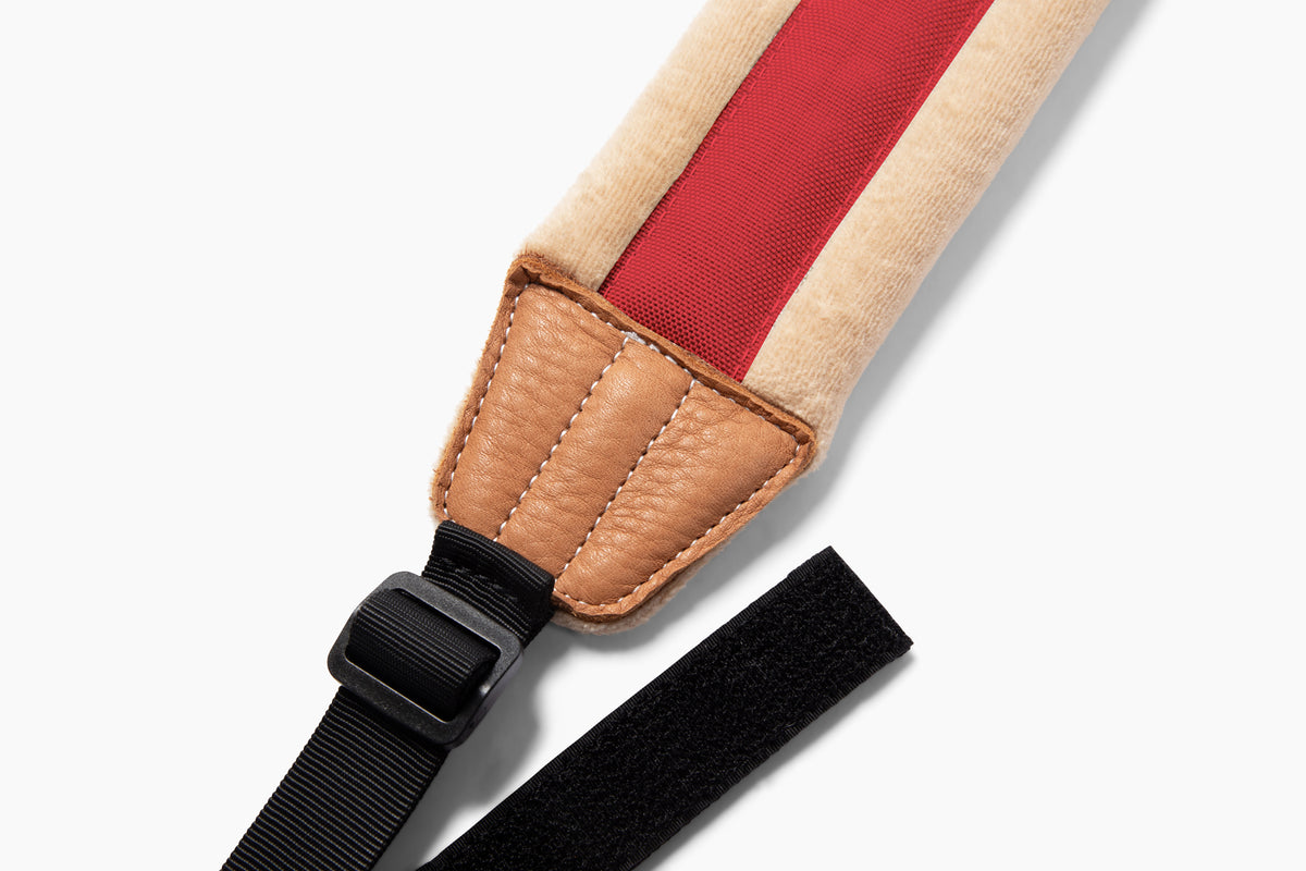 Close-up of a padded shoulder strap with a beige fabric body, a red center stripe, tan leather-like stitching and trim, a black adjustable strap, and a black buckle on a light gray background.