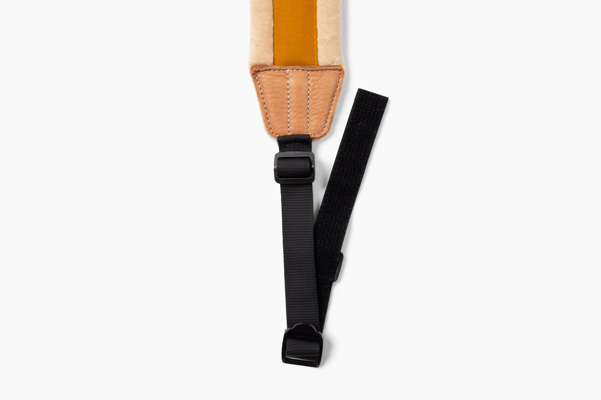 Close-up of a strap accessory with a tan leather patch, cream and mustard yellow fabric section, and a black adjustable strap with plastic buckles, shown on a light gray background.